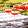 Person pouring a red cocktail from a bottle into two glasses on a striped cloth with a blurred green background.
