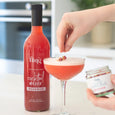 Bottle of 'fling' cocktail mixer Rosebud next to a glass with a red cocktail, with a hand holding a jar of garnish.