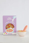 Box of 'tiny harlow' Tiny Loops cereal with a bowl of cereal and spoon on a light gray background