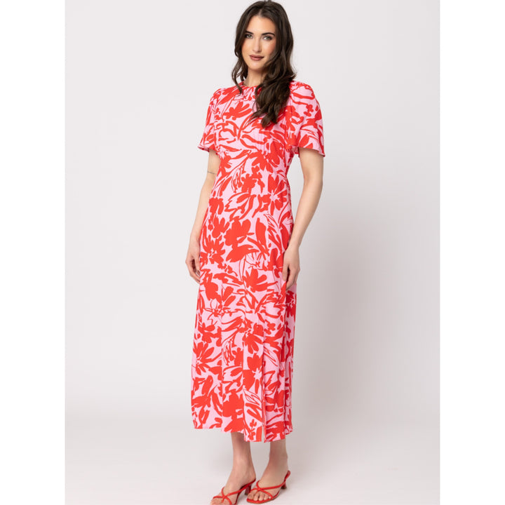 A woman modeling a short-sleeve midi dress with a vibrant coral red and white floral pattern, standing against a light gray studio background.