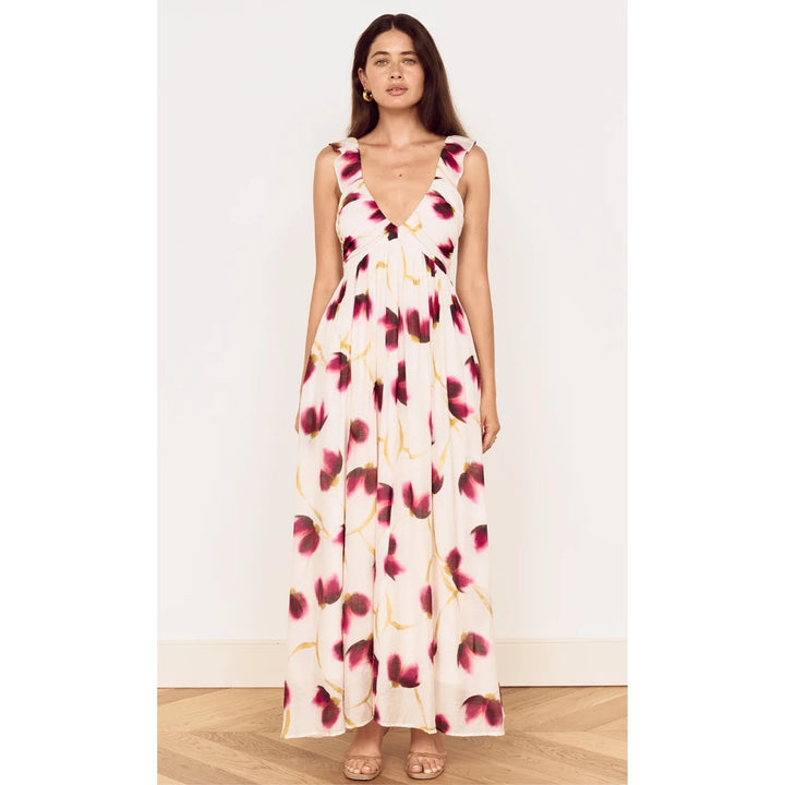A woman modeling a sleeveless, V-neck maxi dress with a cream base and a dark red and green floral print, standing against a plain white wall on a light wood floor.