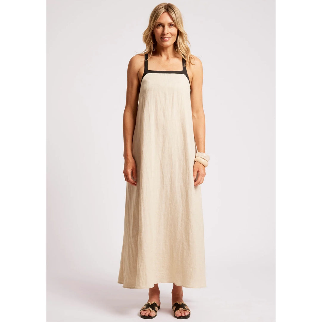 A woman modeling a sleeveless maxi dress in a light beige linen-like fabric with black trim along the square neckline and armholes, paired with black sandals, against a plain light gray studio background.