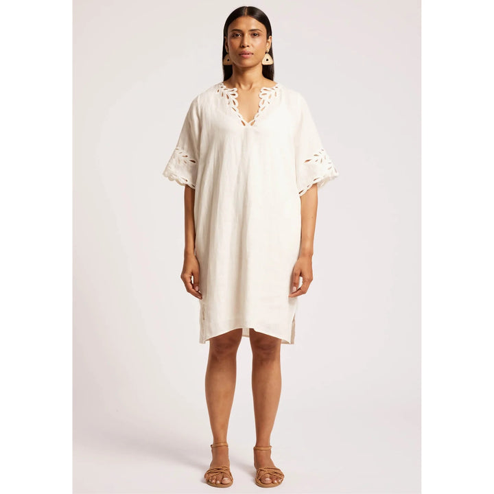A woman modeling a short-sleeve white linen tunic dress with eyelet lace trim at the V-neck and sleeve cuffs, paired with tan leather sandals against a light gray studio background.