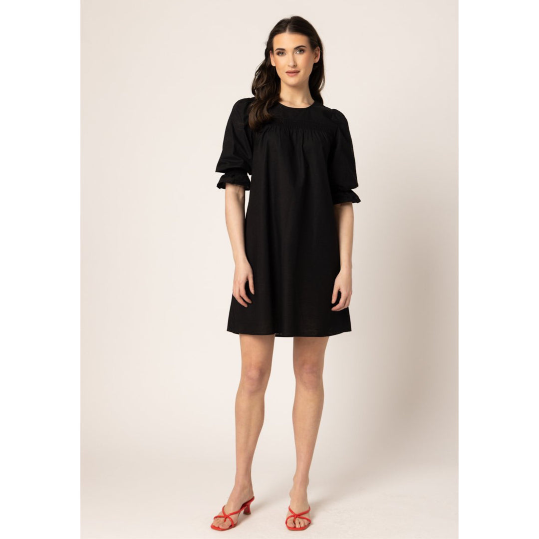 A woman modeling a short black dress with a smocked bodice and ruffled elbow-length sleeves, paired with red heeled sandals, against a plain beige studio background.