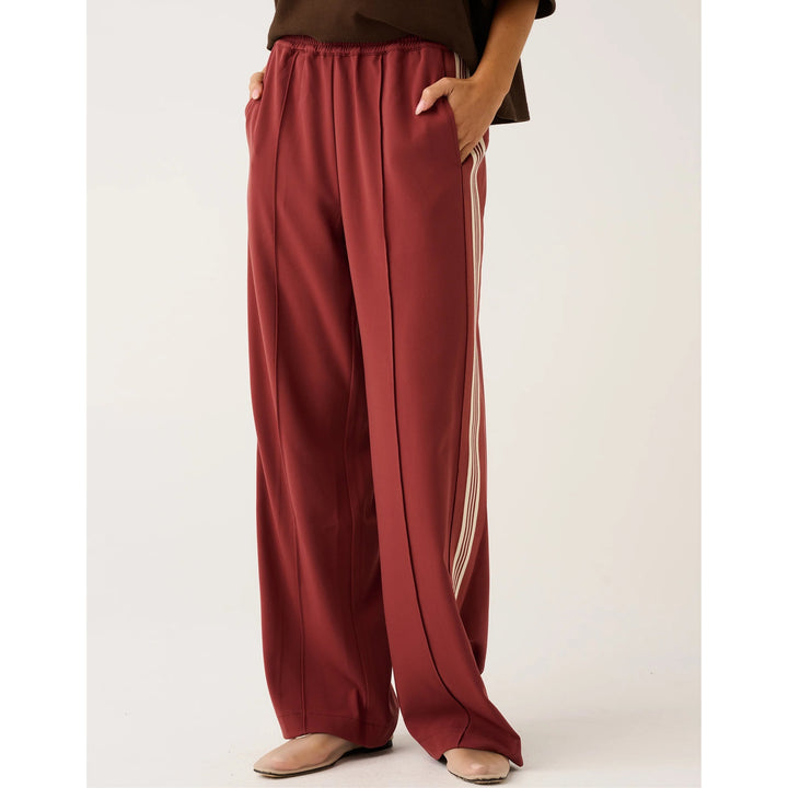 A person wearing wide-leg burgundy pants with a vertical cream stripe down the side, paired with a dark brown top and beige flat shoes, shown against a plain light background.