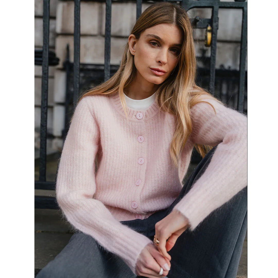 A woman wearing a light pink fuzzy button-front cardigan over a white top and dark gray pants, seated in front of a black metal gate.