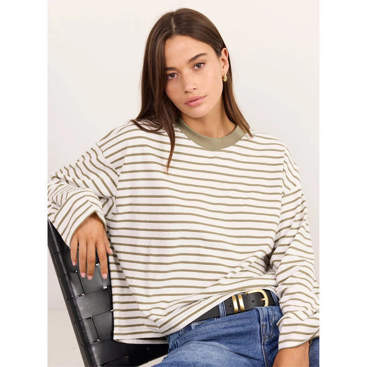 A woman wearing a cream and olive green horizontally striped long-sleeve top, paired with blue jeans and a black belt, sitting on a black woven chair against a light gray background.