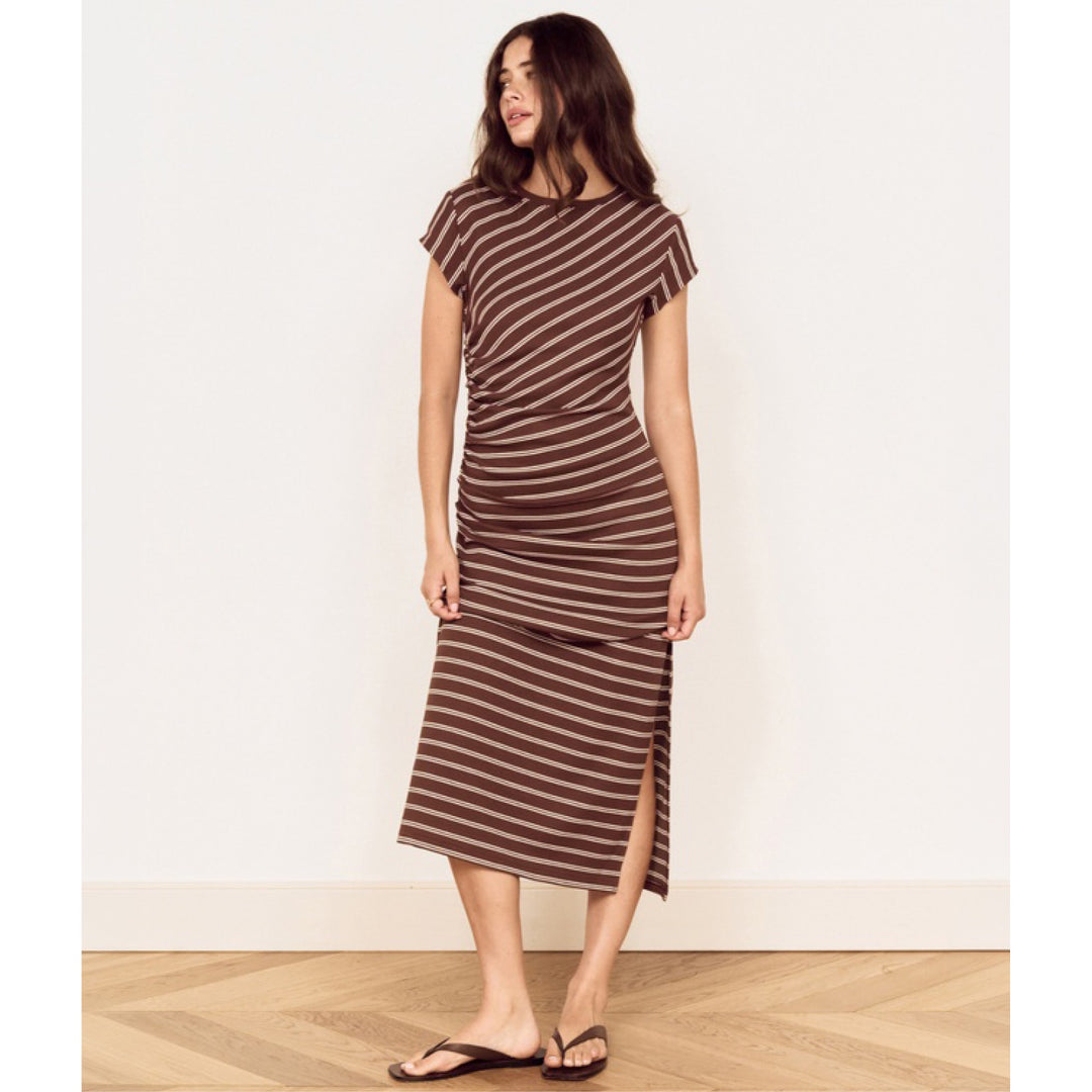 A woman modeling a brown and beige horizontally striped midi dress with a ruched waist and side slit, paired with dark brown flip-flop sandals, standing against a plain off-white wall.