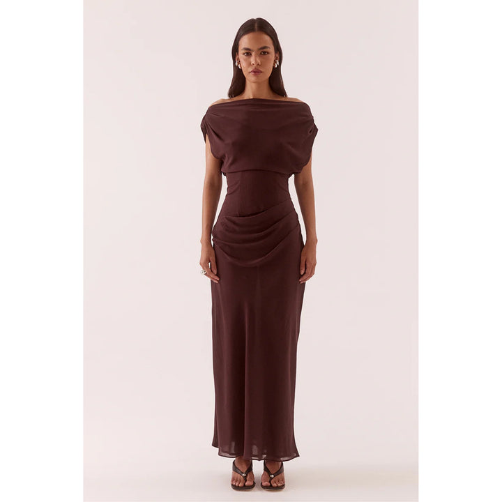 A woman modeling a floor-length off-the-shoulder dress in a deep chocolate brown color, standing against a solid pale pink studio background.