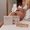 Pregnant woman in a white robe with a pregnancy spa experience kit on a bed.