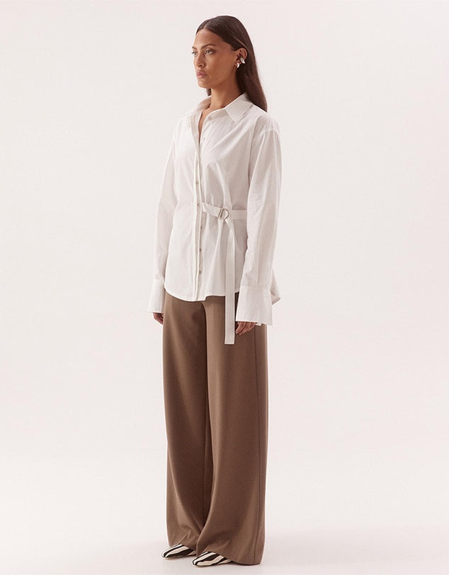 A woman modeling a white long-sleeve button-up shirt with a matching tie belt and wide-leg brown trousers, standing against a plain light background.