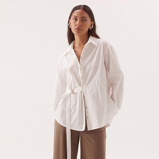 A woman modeling a white long-sleeve button-up shirt with a tie waist belt and brown wide-leg pants against a plain light background.