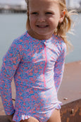 Young girl wearing a floral rash guard and shorts by a body of water.