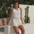 Woman in white sleeveless top and shorts leaning against a white wall with cacti in the background.