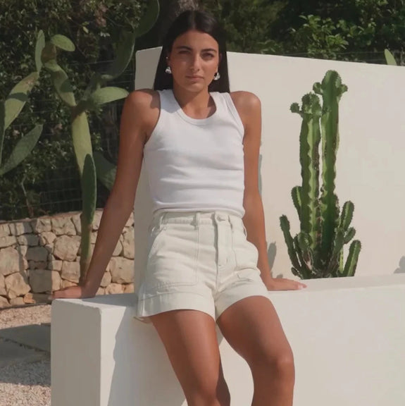 Woman in white sleeveless top and shorts leaning against a white wall with cacti in the background.