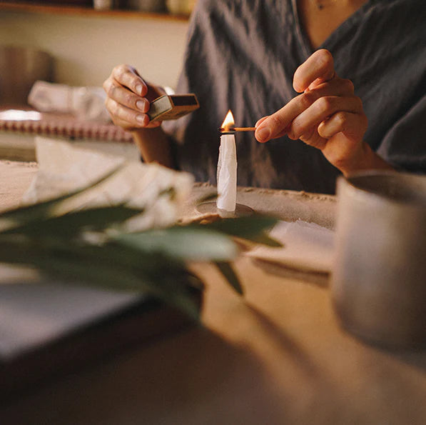Person lighting a candle with a match on a table with plants and a cup.
