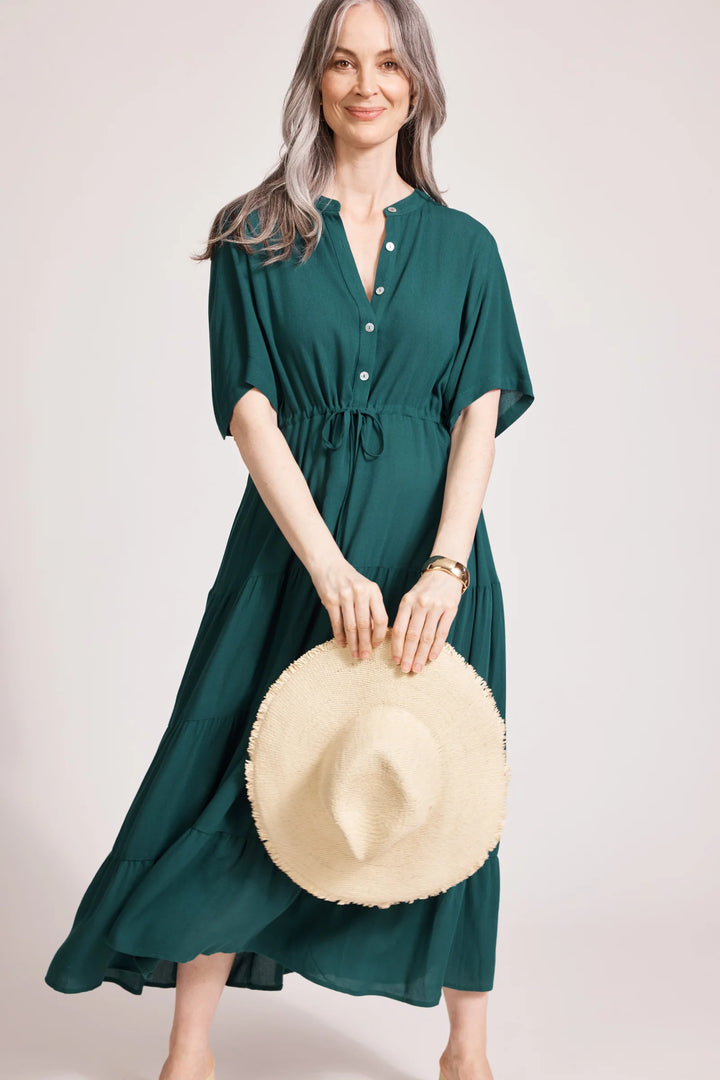 Woman wearing a Eb & Ive green dress holding a beige hat against a light background