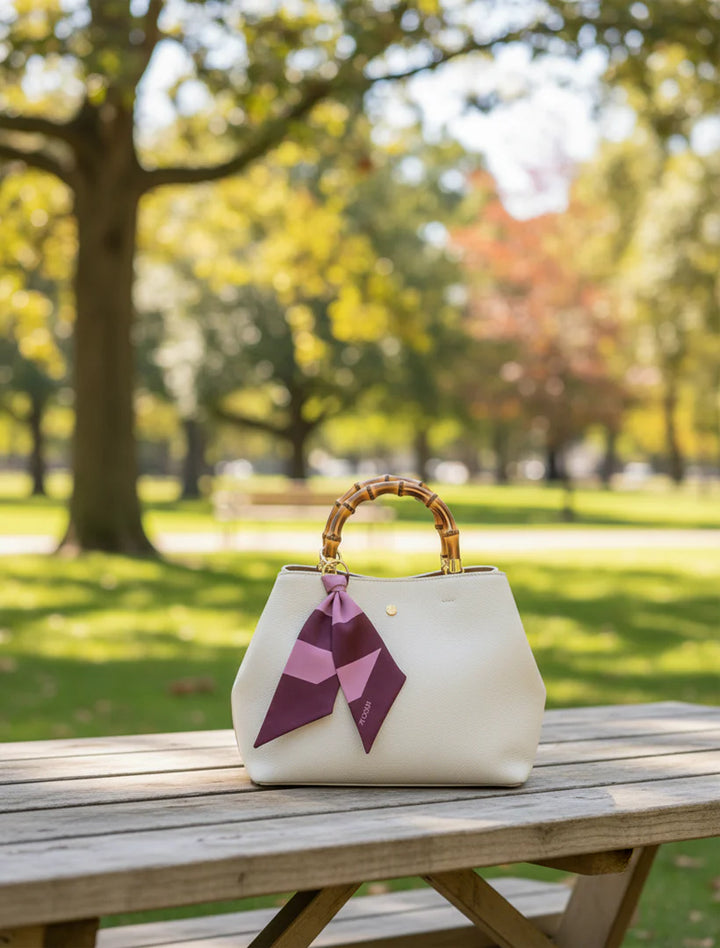White handbag with a bamboo handle and pink accents on a wooden table in a park.