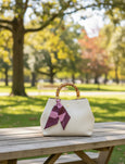 White handbag with a bamboo handle and pink accents on a wooden table in a park.