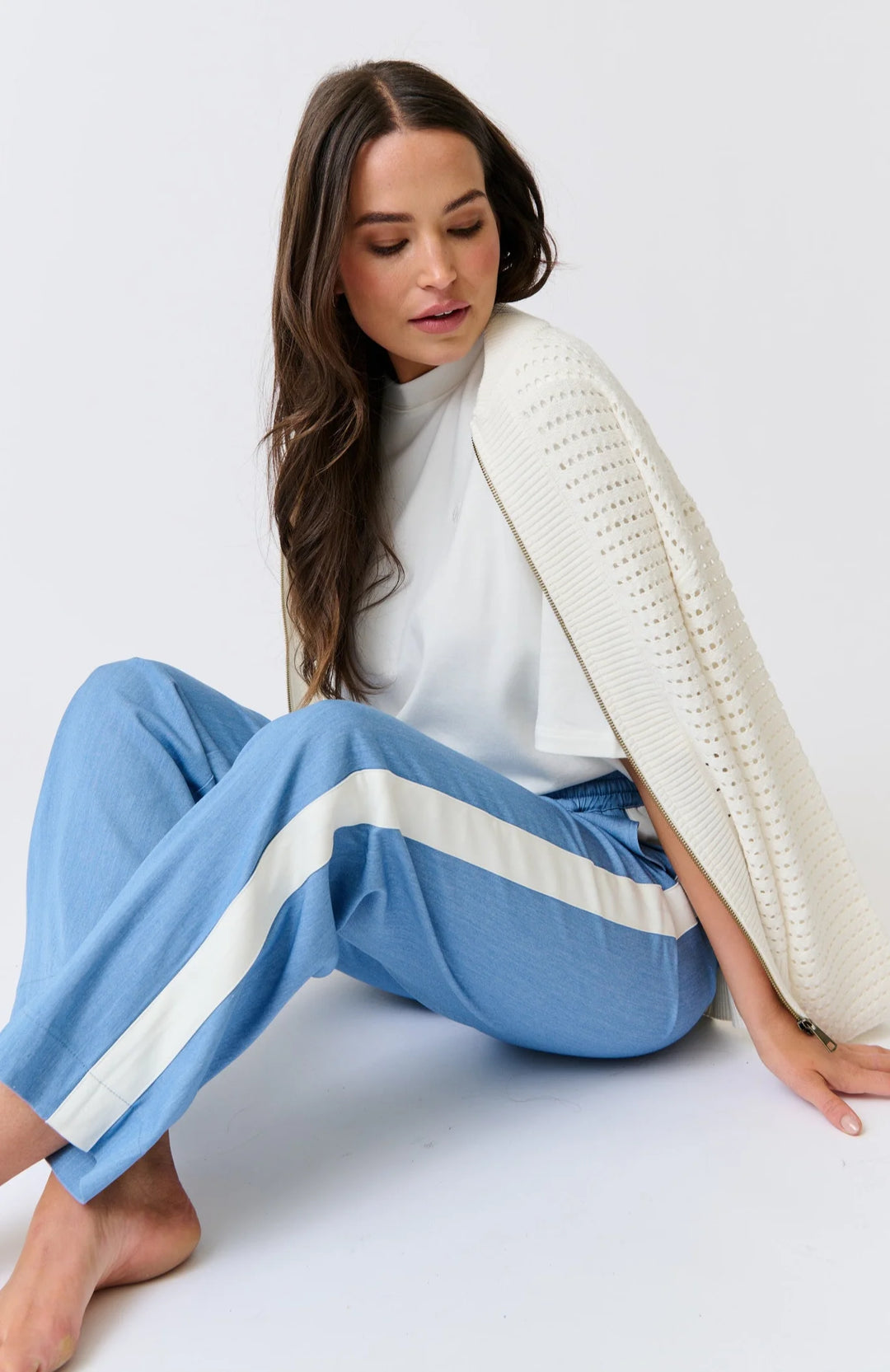 Woman wearing a white sweater and blue pants with a white stripe sitting on a white floor.