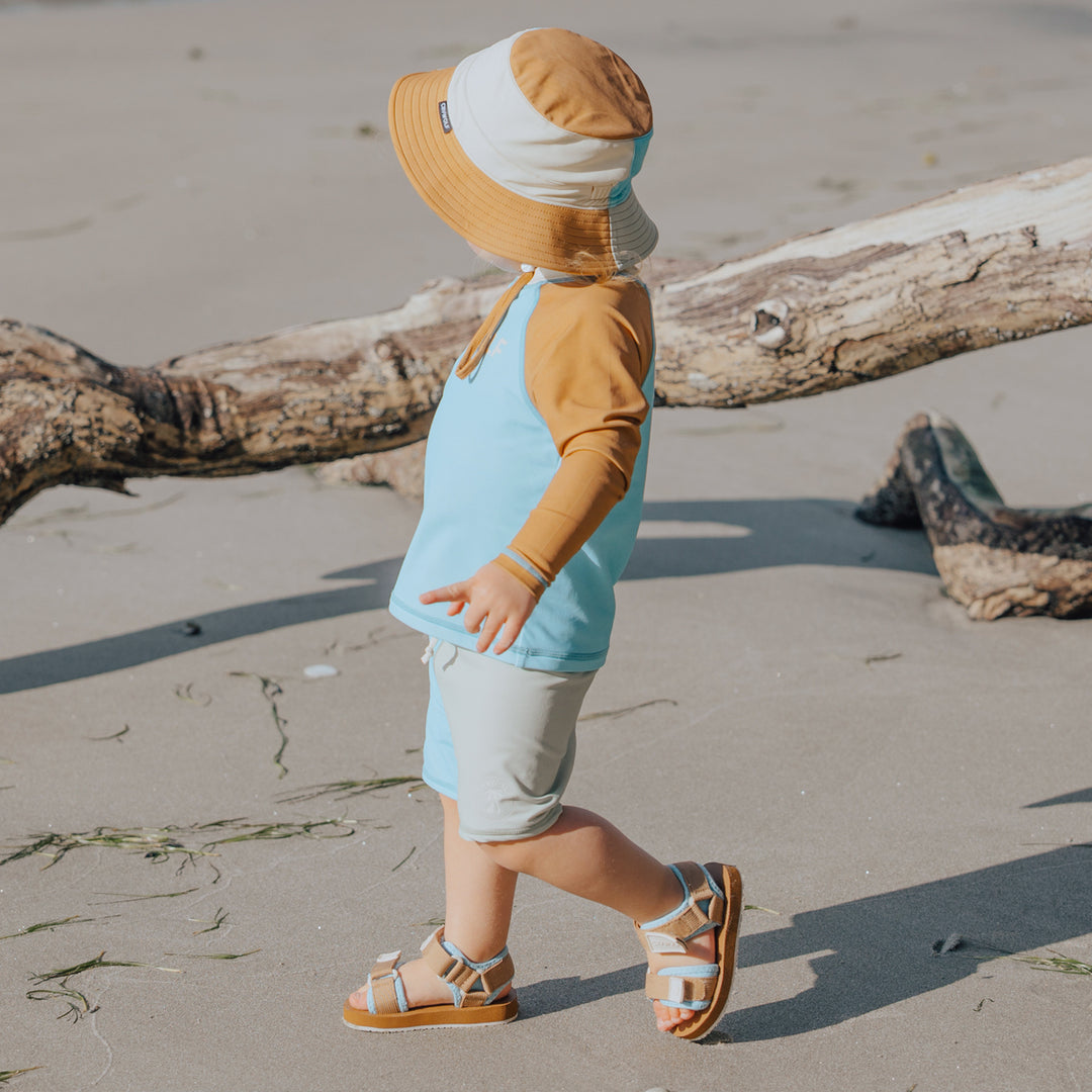 Child wearing a sun hat and colorful crywolf rash vest walking on a sandy beach.