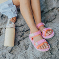 Child's feet wearing crywolf pink beach sandals with orange straps on a sandy surface.