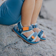 Child wearing blue crywolf beach sandals with orange accents on a sandy surface