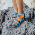 Children's feet wearing crywolf blue sandals with orange accents on a rocky surface.