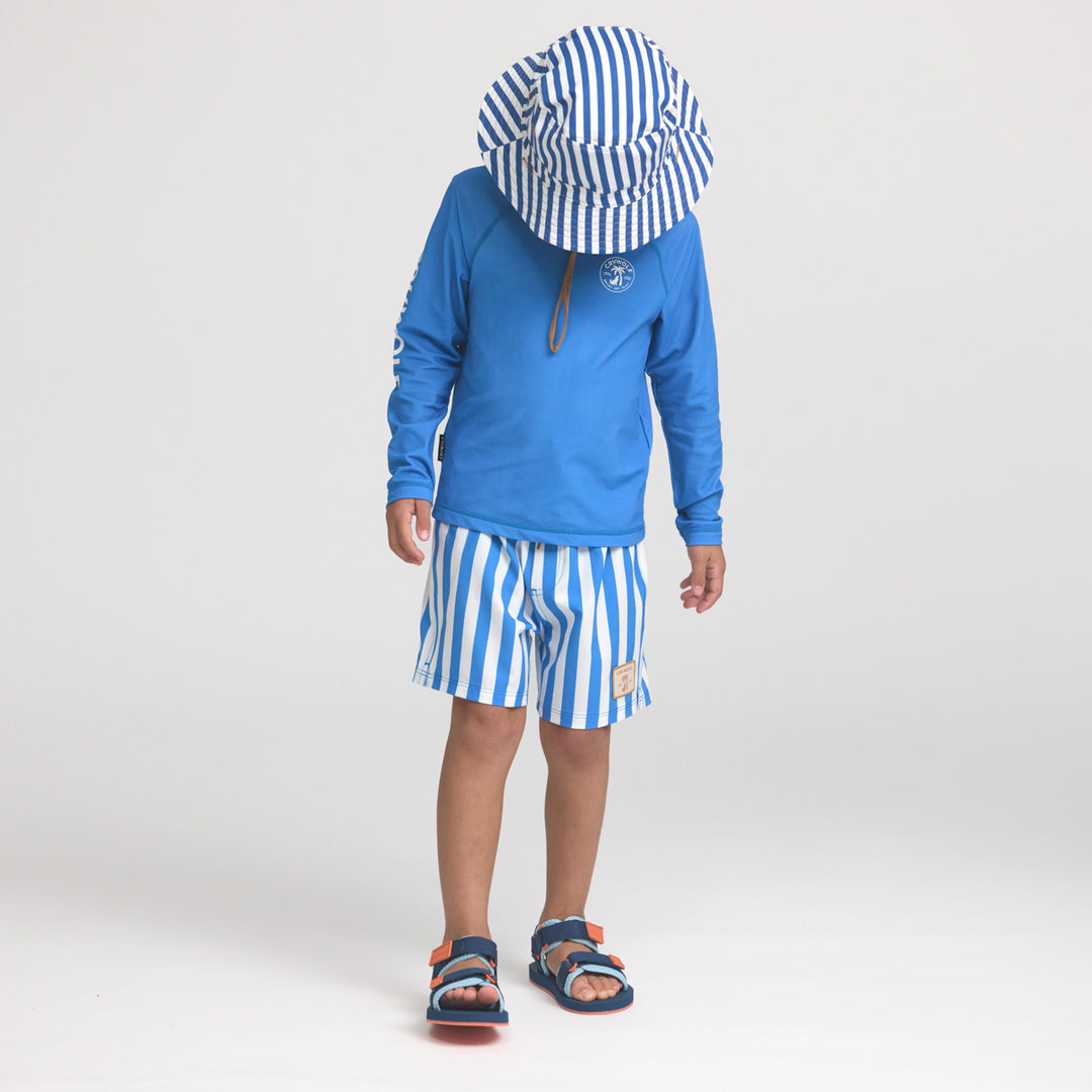 Child wearing a crywolf blue long-sleeve rash vest, striped board shorts, and a matching hat on a white background