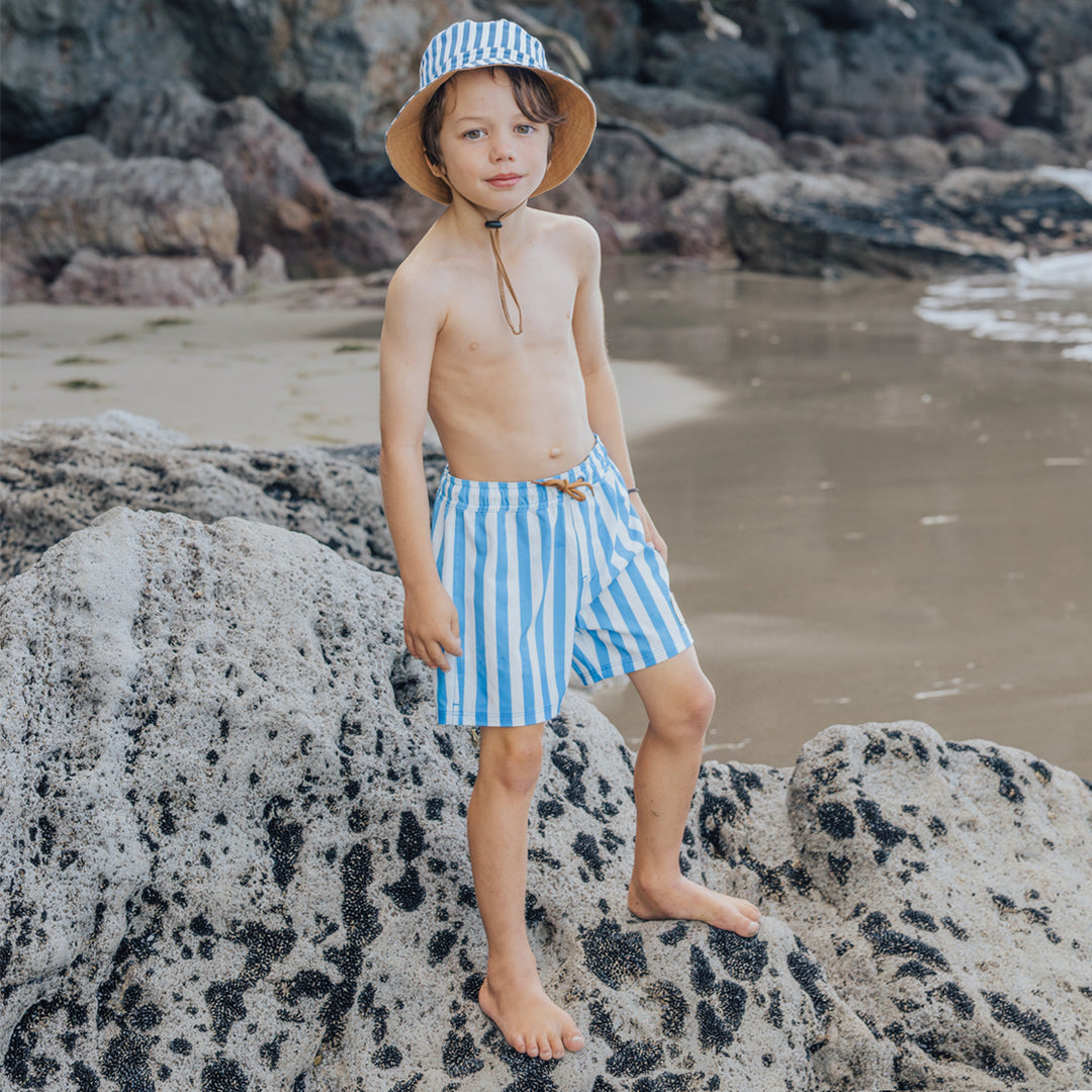 Child wearing crywolf blue and white striped swim shorts and a hat on a rocky beach.