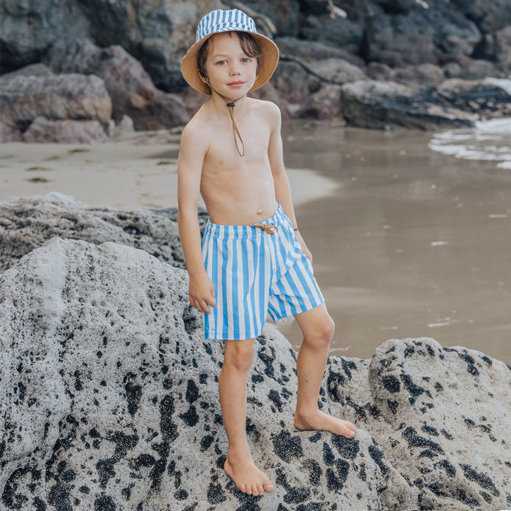Child wearing crywolf blue and white striped swim shorts and a hat on a rocky beach.