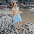 Child wearing crywolf blue and white striped swim shorts and a hat on a rocky beach.