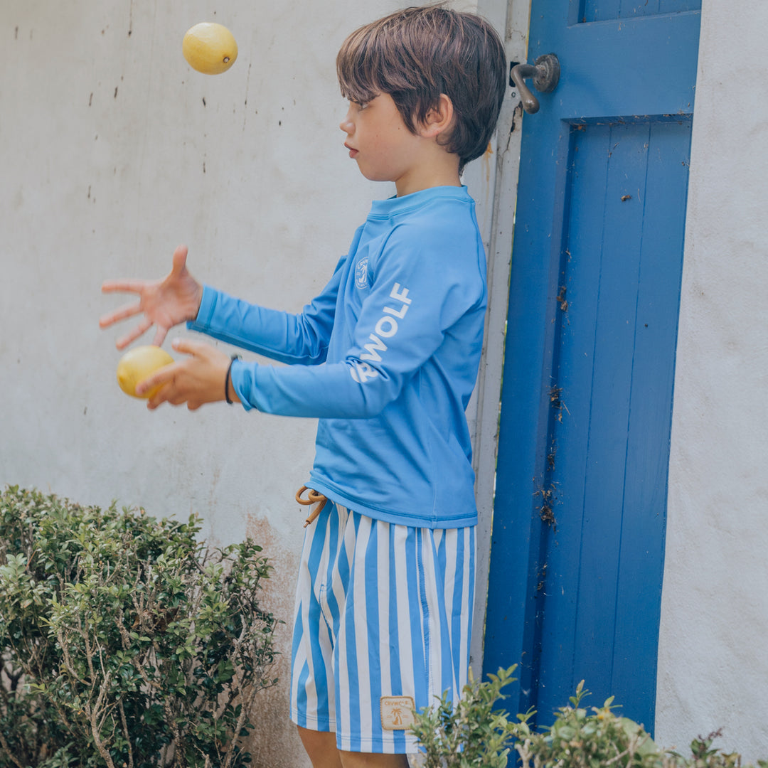 Child in blue rash vest and striped board shorts juggling apples near a blue door.