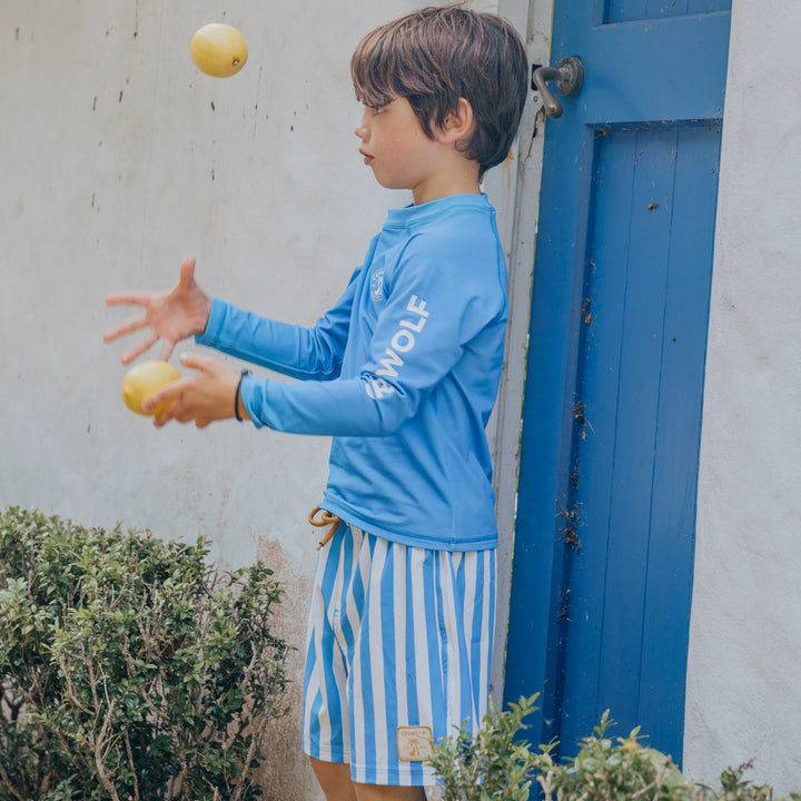 Child in blue rash vest and striped board shorts juggling apples near a blue door.