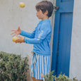 Child in blue rash vest and striped board shorts juggling apples near a blue door.