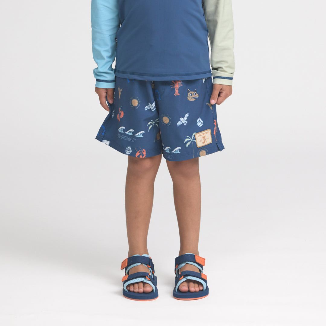 Child wearing a crywolf blue patterned rash vest and board shorts with sandals on a white background