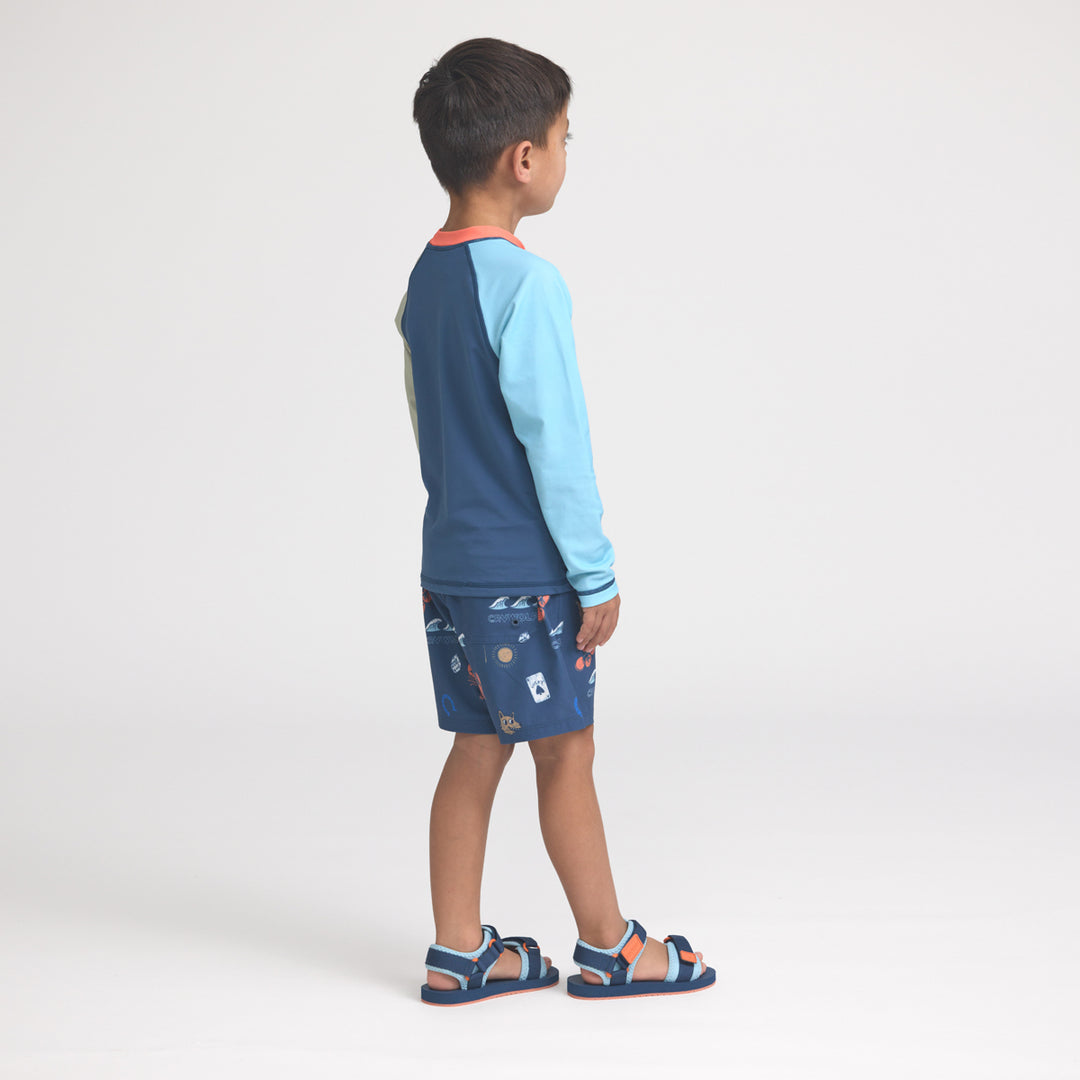 Child wearing a blue and light blue rash vest with patterned board shorts on a white background