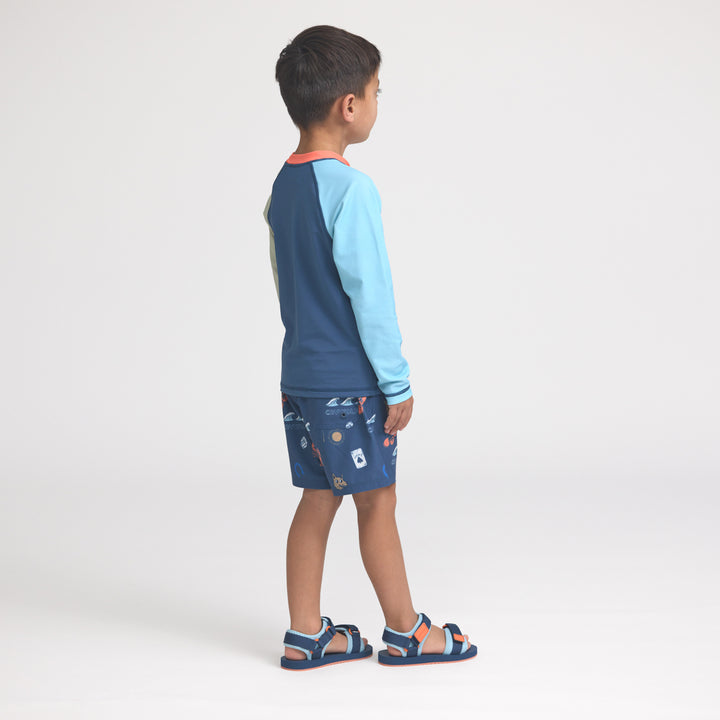 Child wearing a blue and light blue rash vest with patterned board shorts on a white background