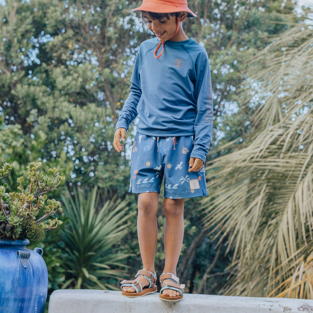 Child wearing a crywolf blue long-sleeve rash vest and  board shorts with a floral pattern, standing outdoors with greenery in the background.
