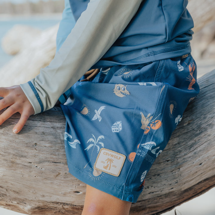 Person wearing blue board shorts with a pattern, standing on a wooden log by a body of water.