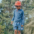 Child wearing a blue long-sleeve rash vest  and patterned shorts with an orange bucket hat outdoors.