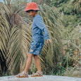 Child wearing a blue long-sleeve shirt and patterned shorts standing outdoors with greenery in the background