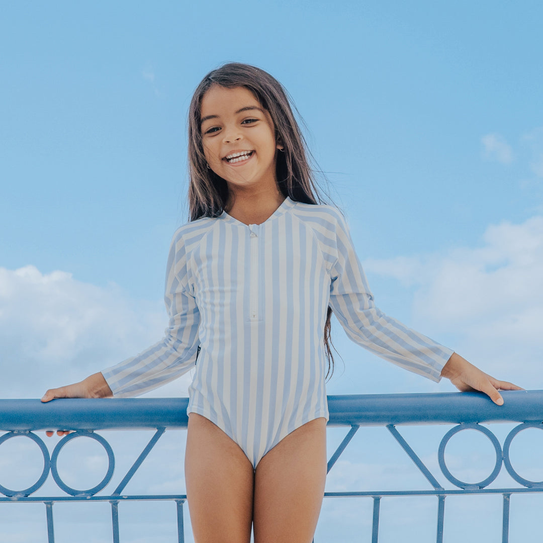 Young girl in a striped crywolf swimsuit standing on a balcony with a blue sky background
