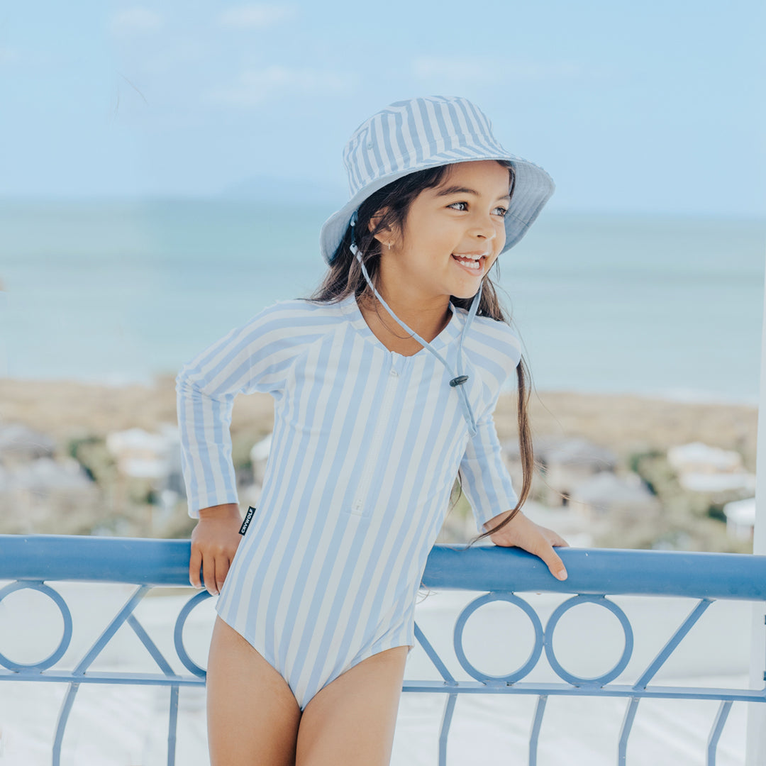 Child wearing a crywolf blue and white striped swimsuit and hat, standing on a balcony with a scenic background.