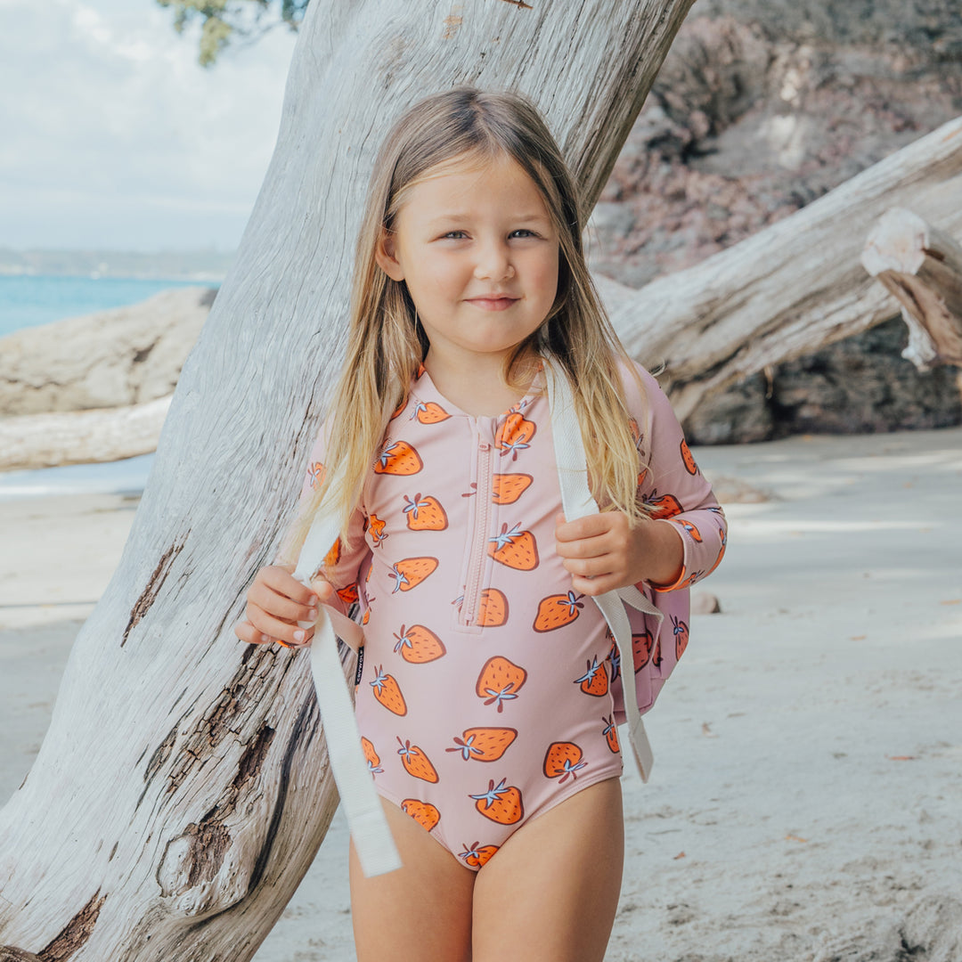 Young girl in a pink crywolf swimsuit with orange strawberry pattern standing by a tree on a beach.