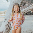 Young girl in a pink crywolf swimsuit with orange strawberry pattern standing by a tree on a beach.