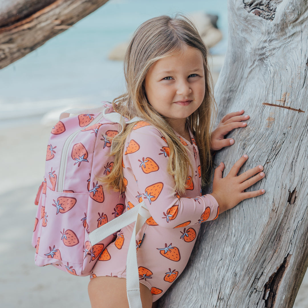 Young girl with a strawberry-patterned crywolf swimsuit and backpack leaning against a tree on a beach.