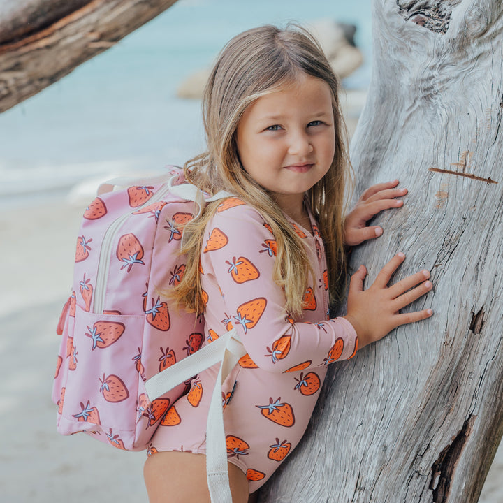 Young girl with a strawberry-patterned crywolf swimsuit and backpack leaning against a tree on a beach.