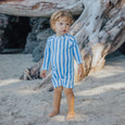 Child wearing a crywolf blue and white striped swimsuit on a sandy beach with driftwood in the background