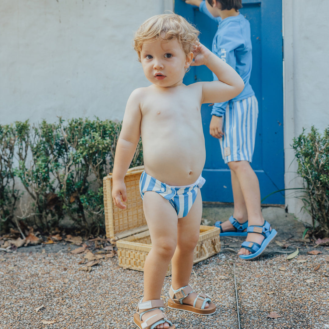 Child wearing a blue and white striped swim nappy outdoors with a wicker basket and another child in the background.